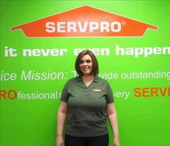 Female employee Heather Lyons standing in front of a green wall with the SERVPRO logo and mission statement below it.