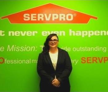 Female employee Sarah Raymond standing in front of a green wall with the SERVPRO logo and mission statement below it.